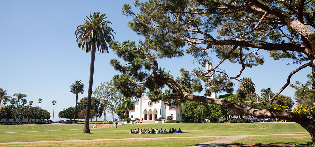 Sacred Heart Chapel from a distance with trees in the foreground and a group of students sitting in a circle on the lawn in front of the chapel.